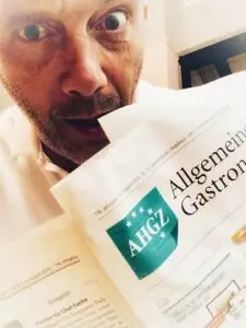 Mann mit überraschter Mine hält eine Zeitung mit dem Titel „Allgemeine Gastronomie“ in der Hand. Hintergrund unscharf.