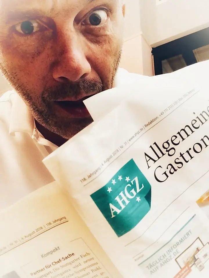 Mann mit überraschter Mine hält eine Zeitung mit dem Titel „Allgemeine Gastronomie“ in der Hand. Hintergrund unscharf.