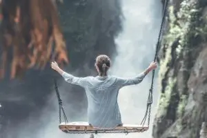 Frau auf einer Holzschaukel, Blick auf einen Wasserfall, umgeben von grünen Felsen und Nebel. Entspannte Atmosphäre.
