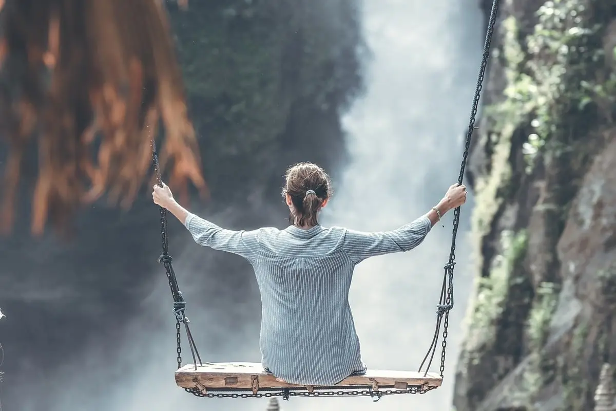 Frau auf einer Holzschaukel, Blick auf einen Wasserfall, umgeben von grünen Felsen und Nebel. Entspannte Atmosphäre.