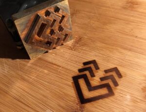 Stempel aus Metall mit geometrischem Muster, daneben der Abdruck auf einem hellen Holzuntergrund.