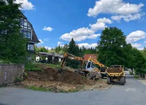 Baustelle mit zwei Baggern und einem Muldenkipper auf einer unebenen Erde, umgeben von Bäumen und Wohnhäusern.