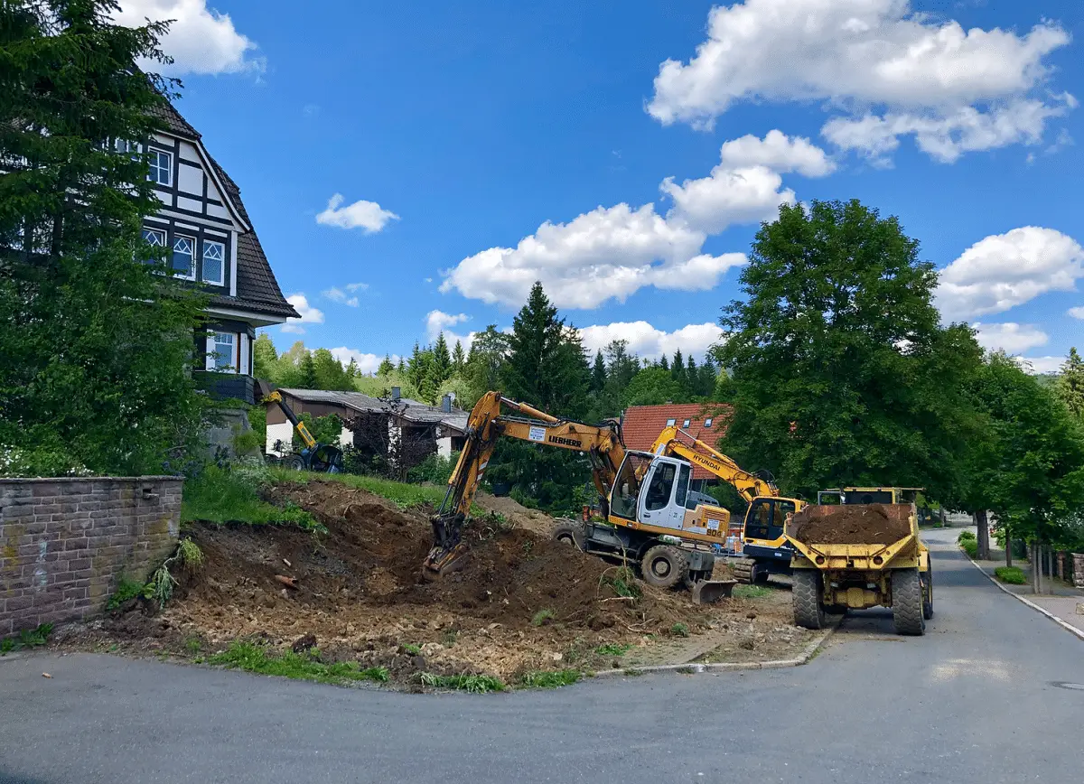 Baustelle mit zwei Baggern und einem Muldenkipper auf einer unebenen Erde, umgeben von Bäumen und Wohnhäusern.