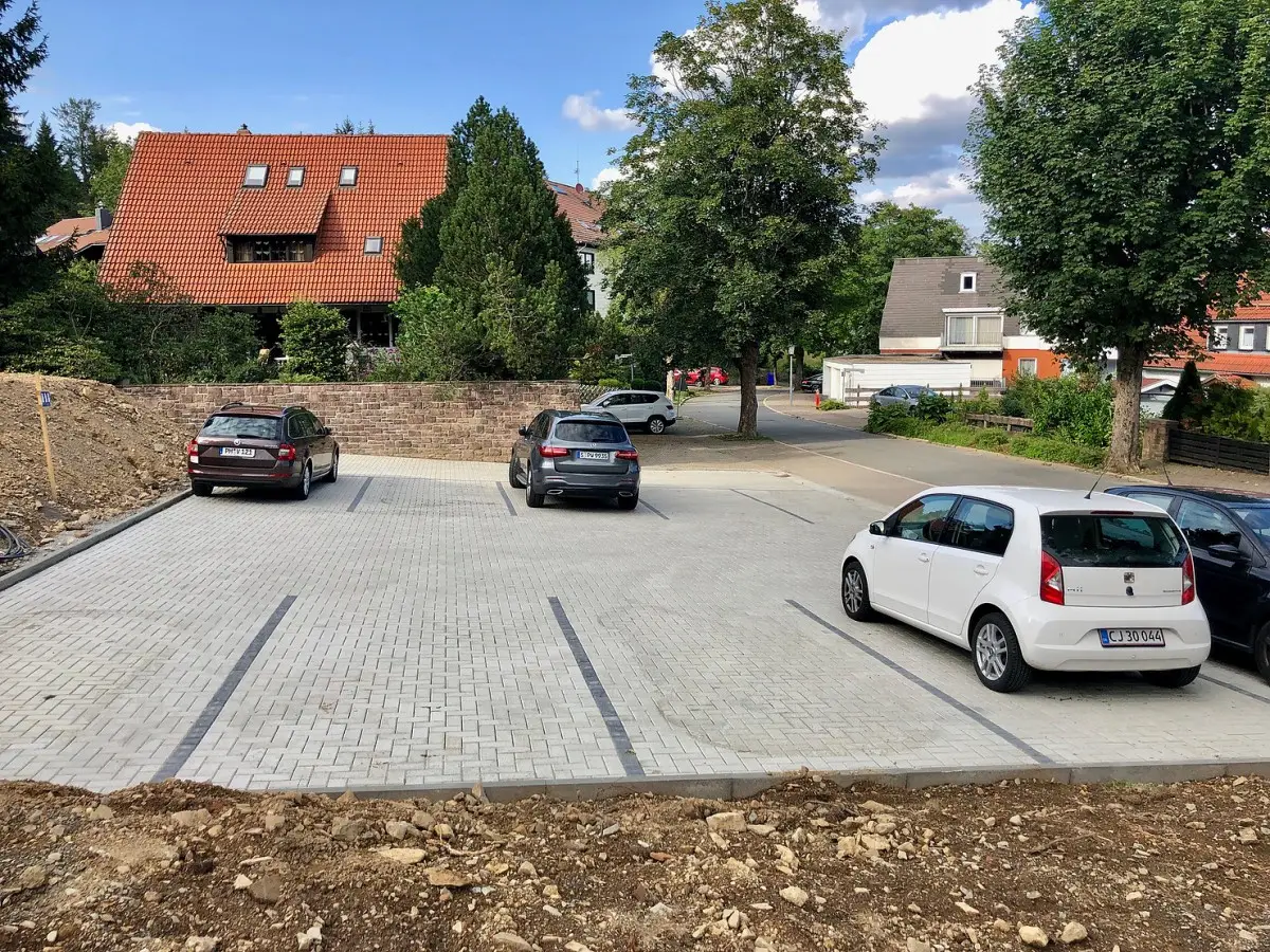 Parkplatz mit vier Autos auf neu gepflasterter Fläche, im Hintergrund Wohnhäuser und eine Baustelle.