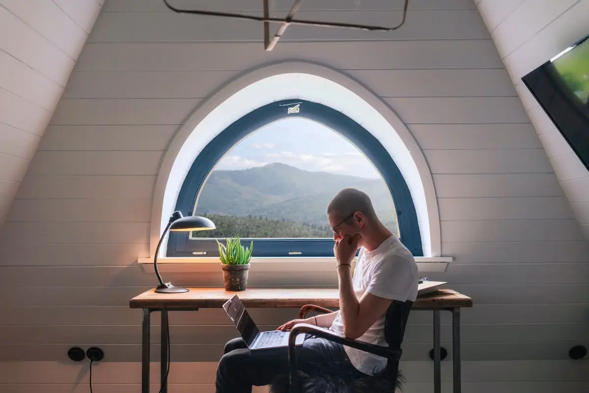 Mann mit Brille sitzt am Schreibtisch, arbeitet am Laptop und blickt nachdenklich aus einem großen Fenster auf Berge.