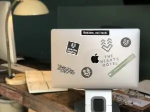 Laptop mit Aufklebern der Hearts Hotel und Together auf einem Holzschreibtisch, mit einer Lampe und einem Planer im Hintergrund.