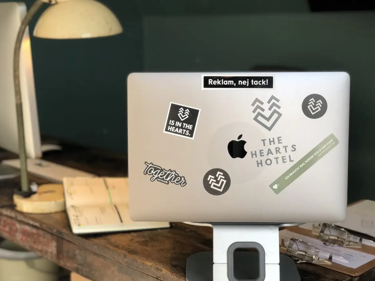 Laptop mit Aufklebern der Hearts Hotel und Together auf einem Holzschreibtisch, mit einer Lampe und einem Planer im Hintergrund.