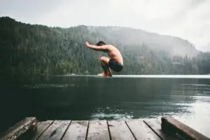Junger Mann springt von einem Holzsteg in einen klaren See, umgeben von einer bewaldeten Landschaft.