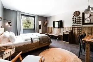 Rustikales Hotelzimmer mit Holzmöbeln, Doppelbett, Schreibtisch, Wanddekoration, Fenster mit Waldblick und Fernseher.