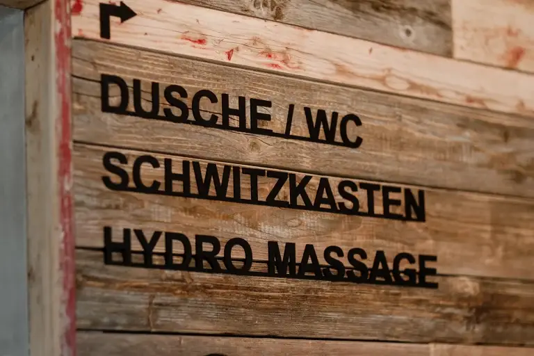 ALT-Text: Wegweiser aus Holz mit Beschriftungen für Dusche/WC, Schwitzkasten und Hydro Massage.