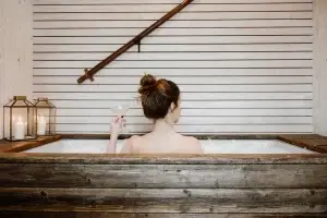 Frau mit hochgestecktem Haar sitzt in einer Badewanne, hält ein Sektglas und blickt auf eine Holzverkleidung. Kerzen sind sichtbar.