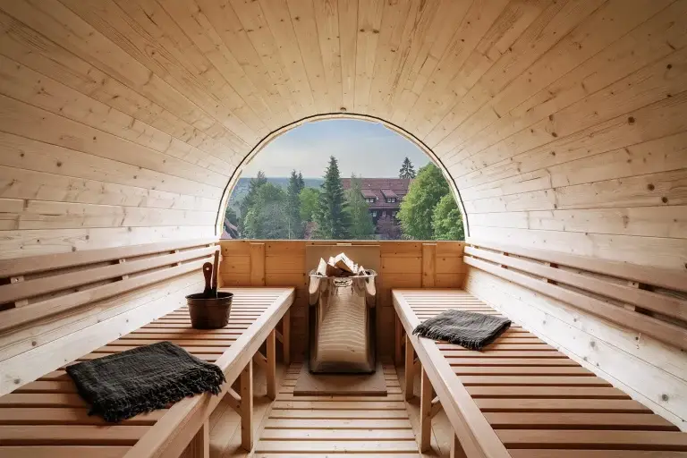 Sauna mit Holzbänken, Blick auf Bäume und Gebäude durch große Fenster; im Raum sind ein Eimer und ein Holzofen sichtbar.