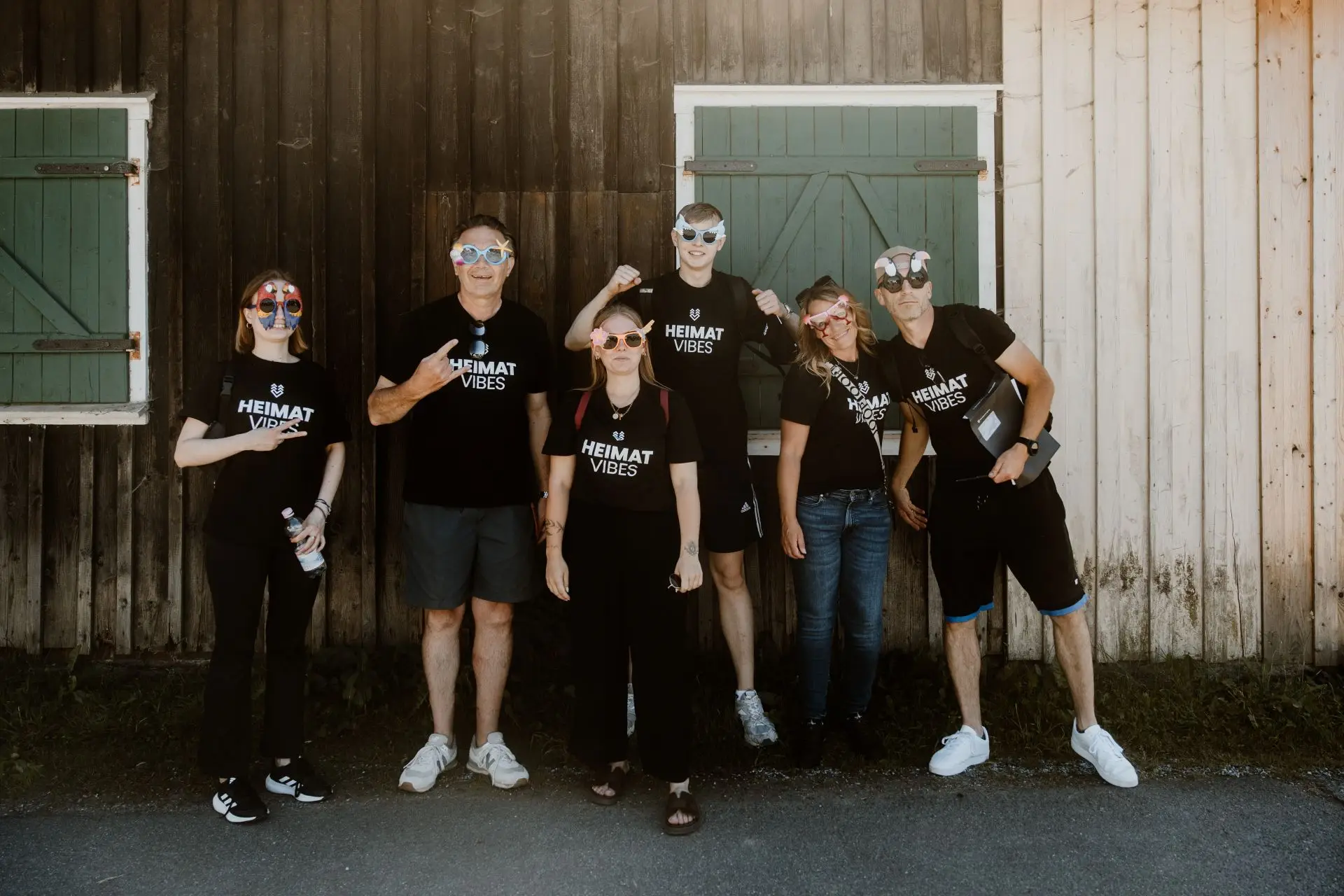 Sechs Personen in Heimat Vibes-T-Shirts stehen lächelnd vor einem Gebäude mit grünen Fensterläden und tragen bunte Brillen.