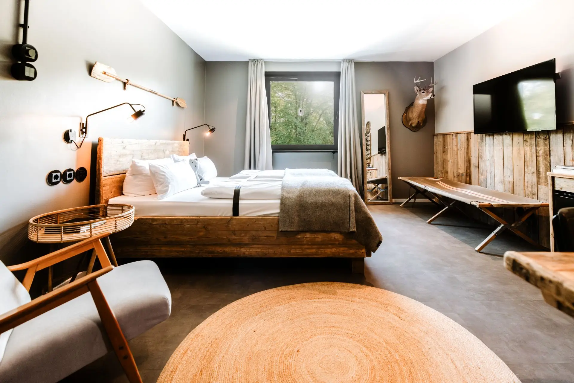 Helles Hotelzimmer mit Doppelbett, Holzdetails, stimmungsvollem Licht und Blick auf Bäume durch das Fenster.