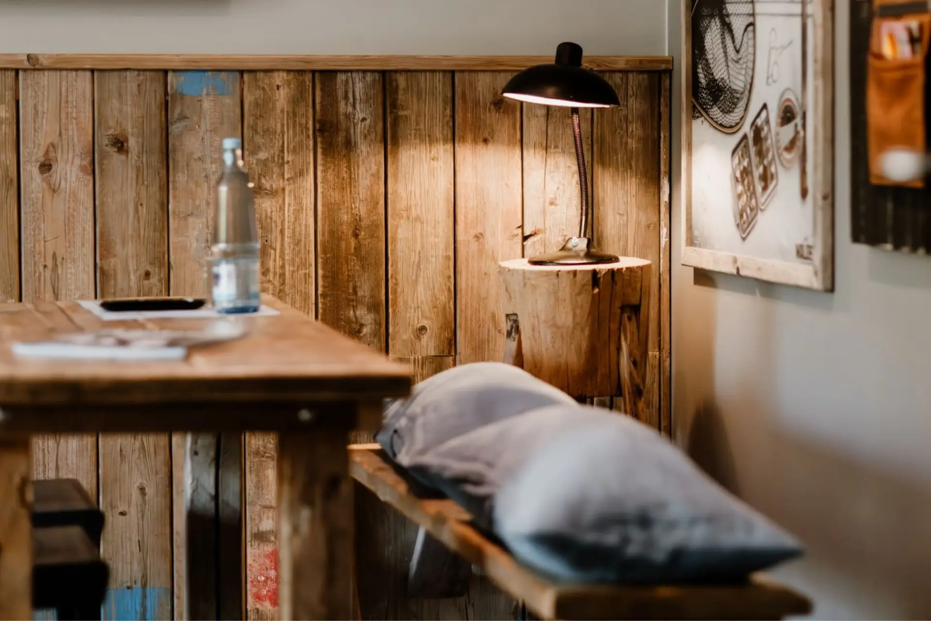 Holztisch mit Wasserglas, Kissen auf einer Bank und einer Tischlampe an der Wand, rustikales Ambiente mit Holzverkleidung.