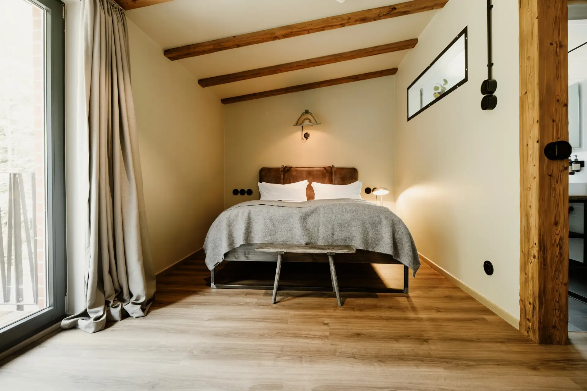 Helles Schlafzimmer mit Holzbalkendecke, Doppelbett, einem Holztisch und großen Fenstern. Beige Wände und Holzfußboden.
