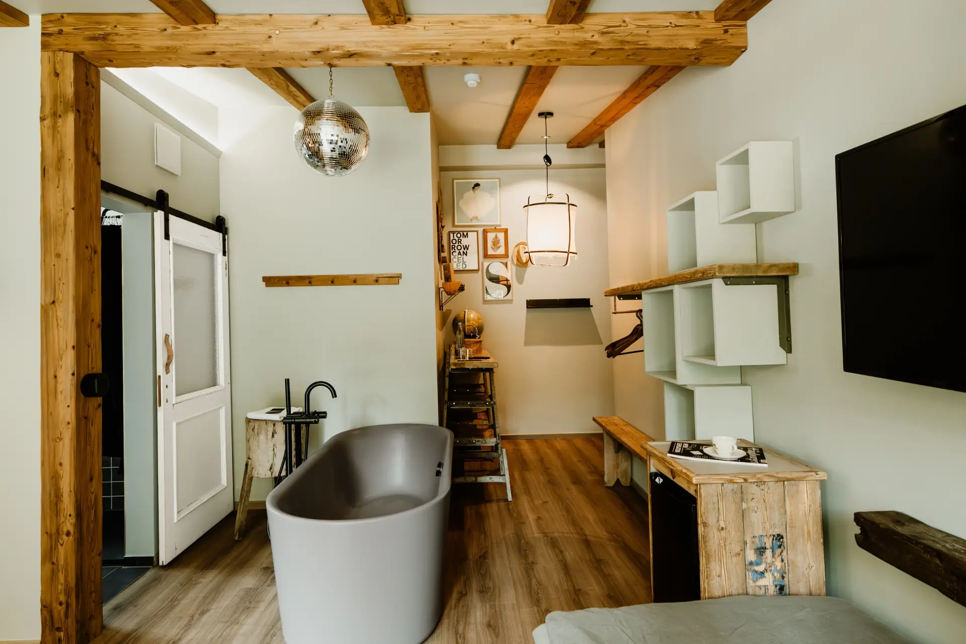 Modernes Badezimmer mit freistehender Badewanne, Holzregalen, Spiegelkugel und TV. Helle Farben und stilvolle Einrichtung.