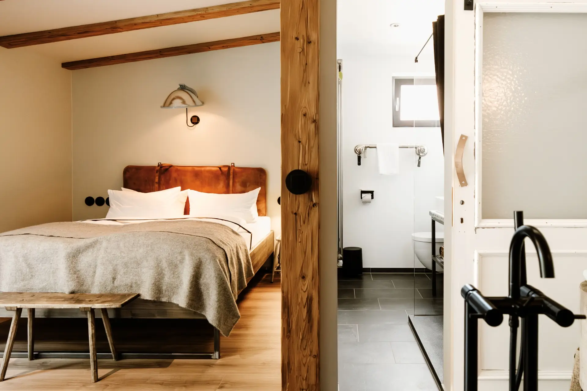 Bild zeigt ein modernes Hotelzimmer mit Doppelbett, Holzbank und Badezimmer mit Dusche und WC. Es ist hell und stilvoll eingerichtet.