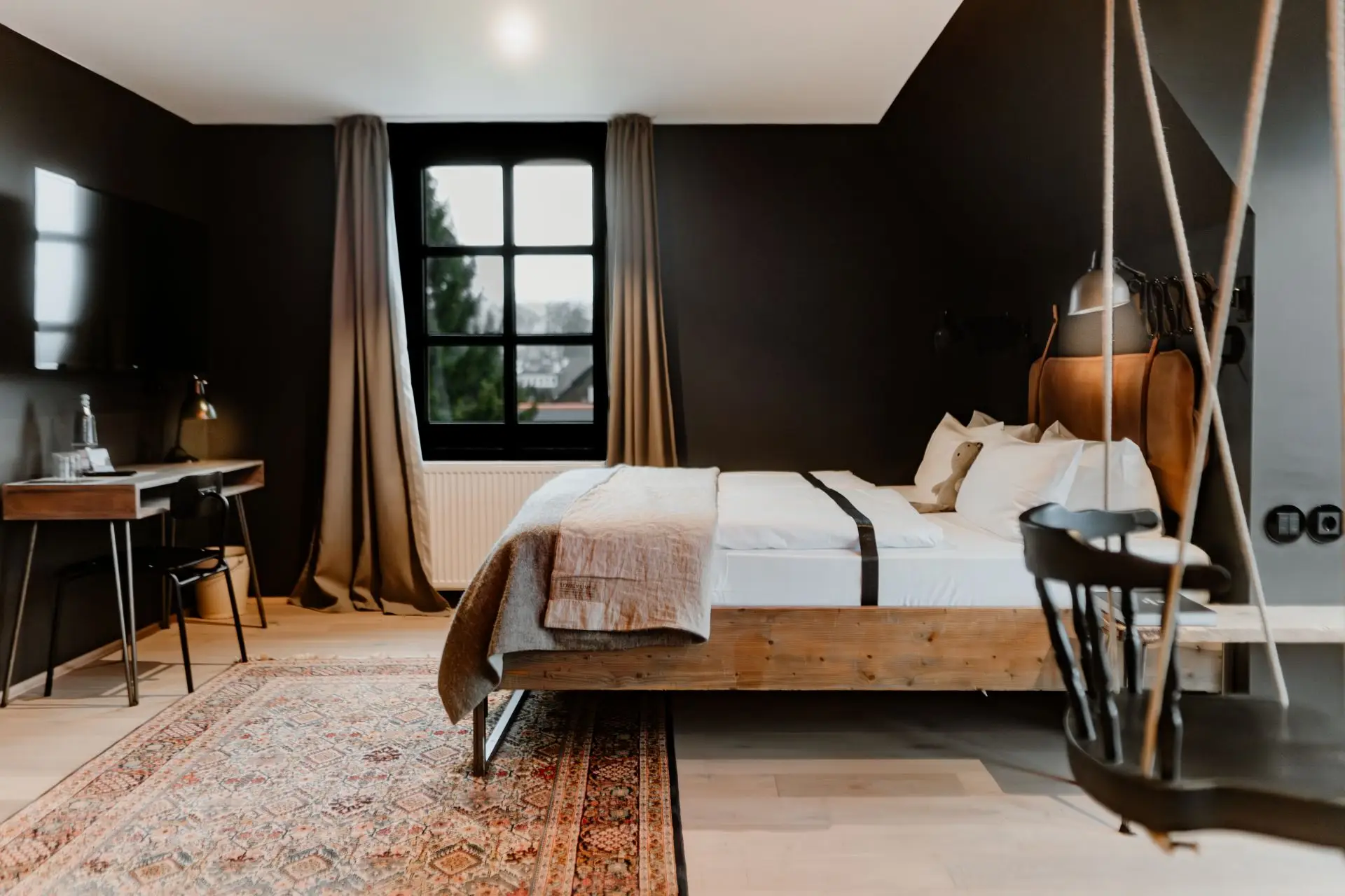 Moderne Hotelzimmer mit Holzherd, Schreibtisch, Fernseher und Hängesessel. Fenster mit Blick ins Freie. neutrale Farbpalette.