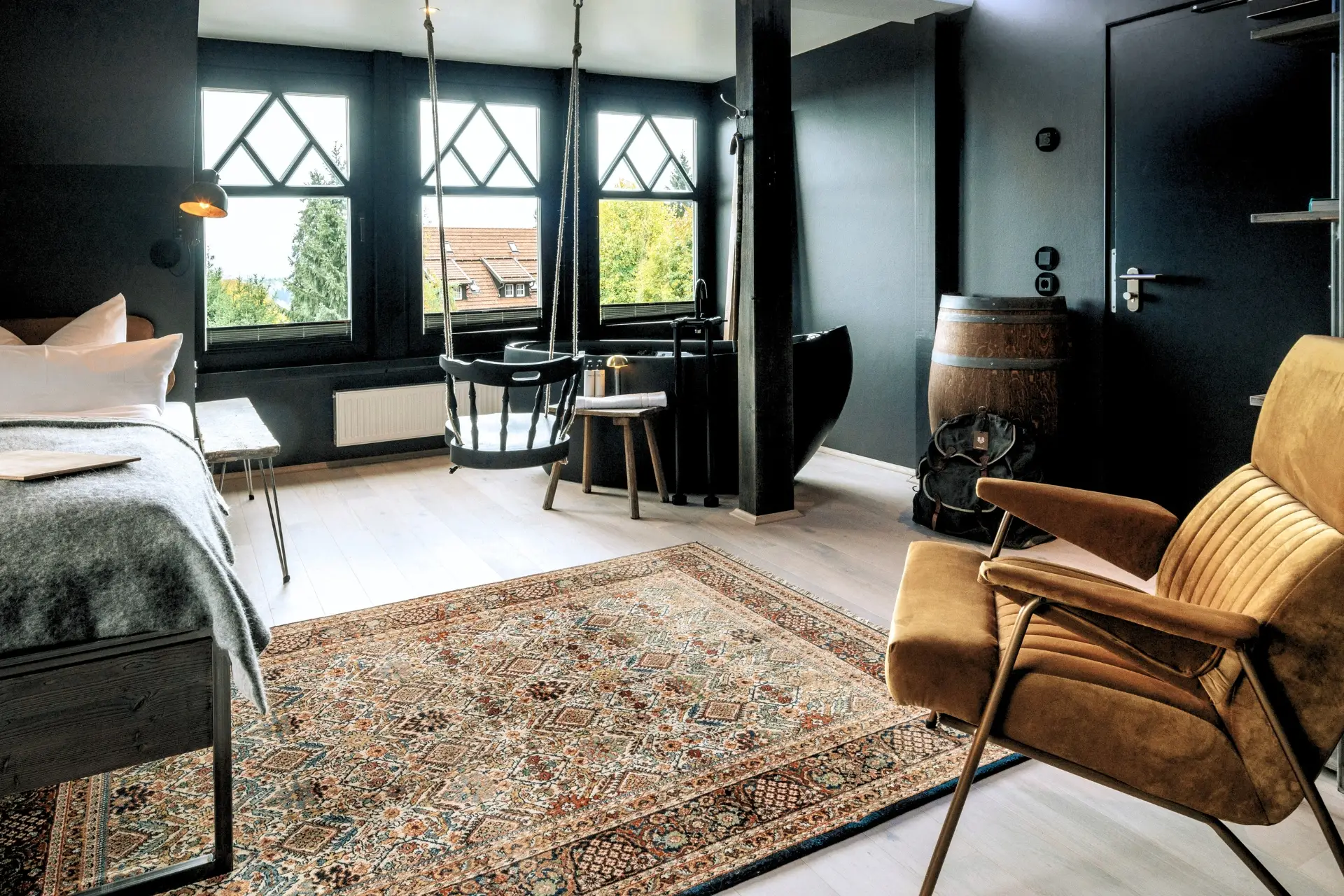 Modernes Zimmer mit Holzfußboden, einem Bett, einer Schaukel, einer Sitzecke und Blick auf die Natur durch große Fenster.