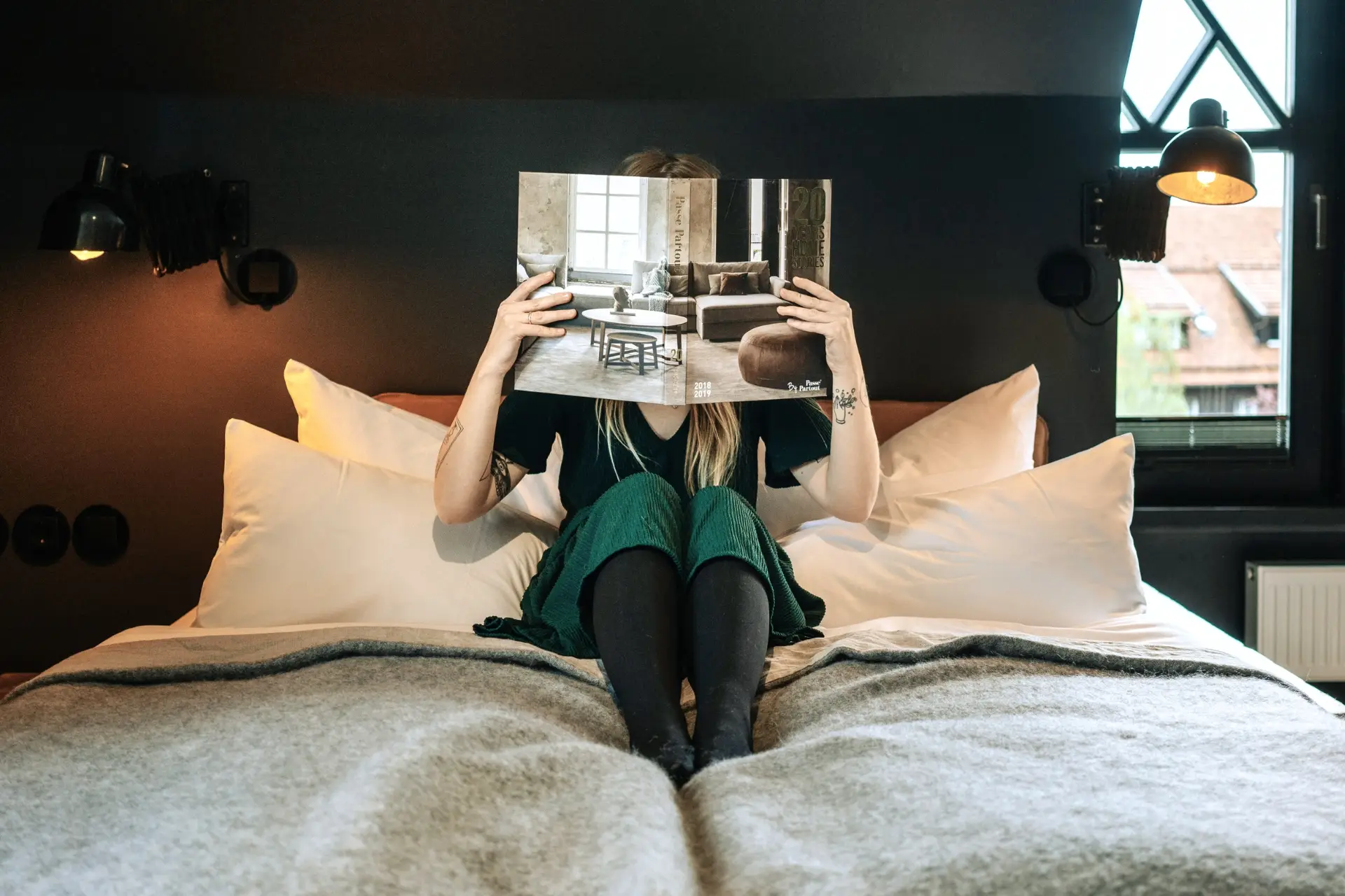 Frau sitzt auf einem Bett, hält ein Magazin über Inneneinrichtung und blickt darauf. Der Raum ist modern und gemütlich.