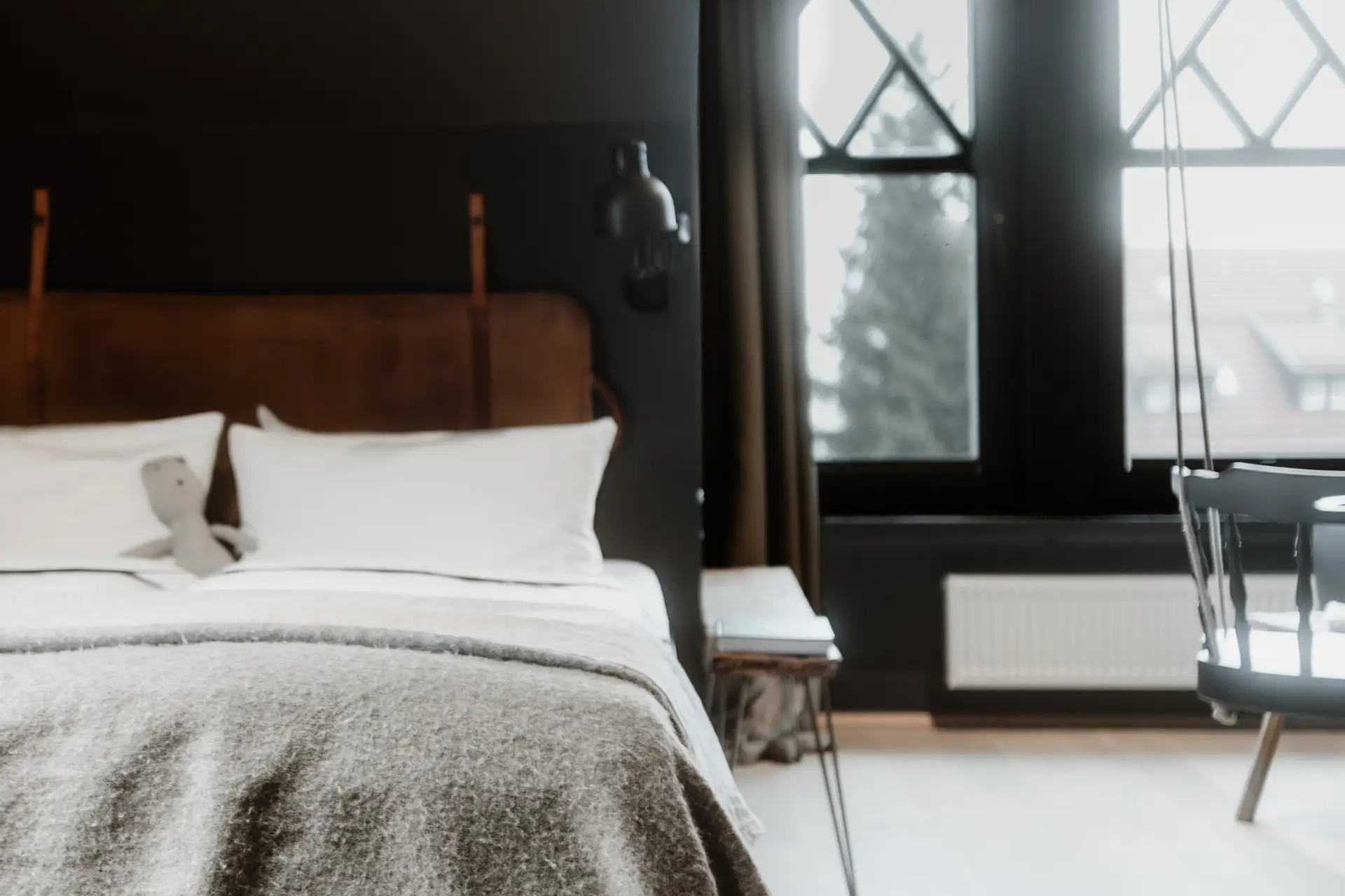 Schlafzimmer mit einem Bett, grauer Decke und einem Stofftier. Fenster mit Blick auf einen Baum und Stuhl im Hintergrund.