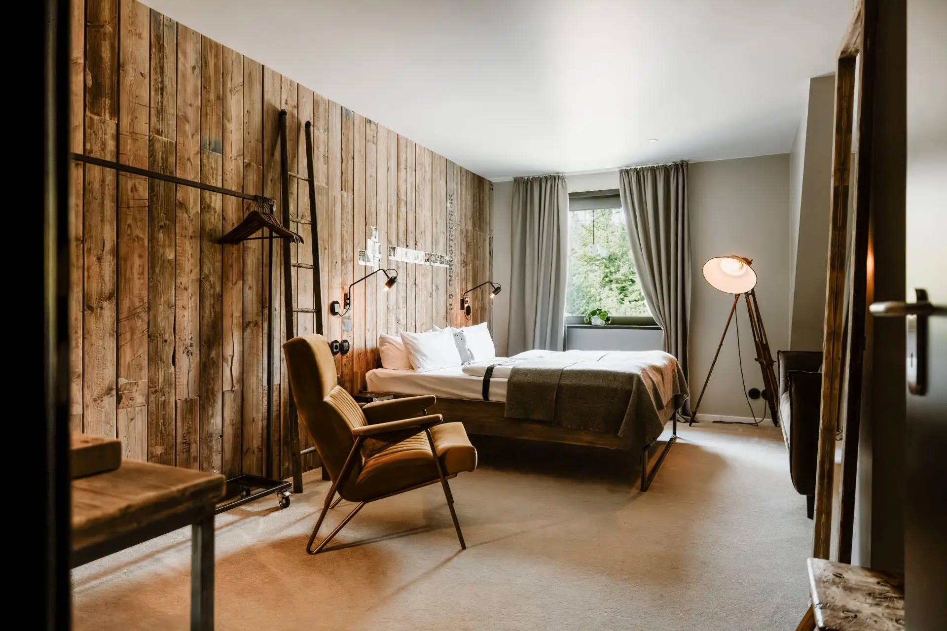 Helles Hotelzimmer mit Holzwänden, Doppelbett, beigem Sessel, Kleiderständer und Fensterblick ins Freie.