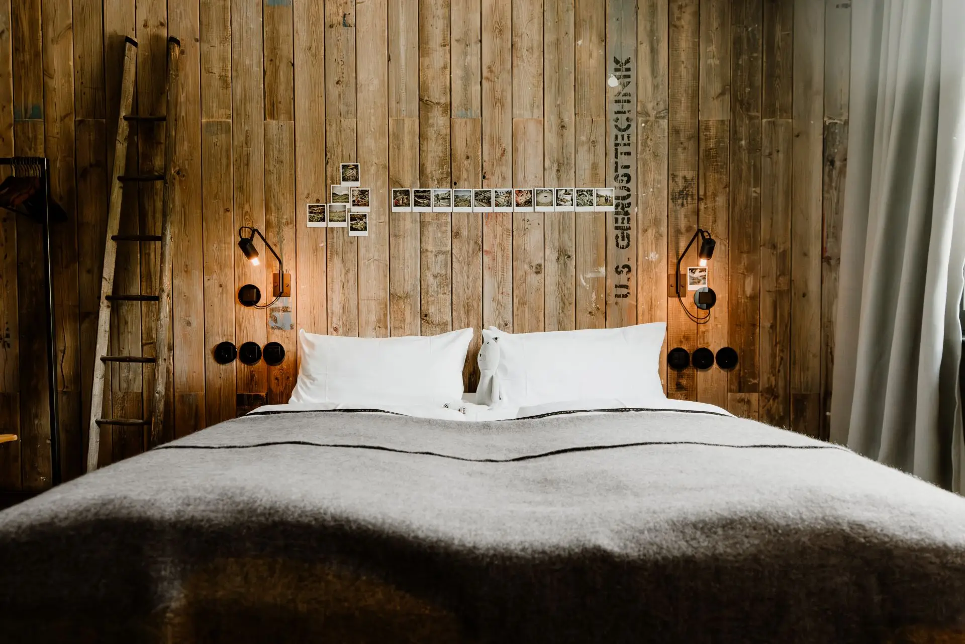 Gemütliches Schlafzimmer mit Holzvertäfelung, Doppelbett und Polaroid-Fotos an der Wand. Eine Leiter steht links neben dem Bett.