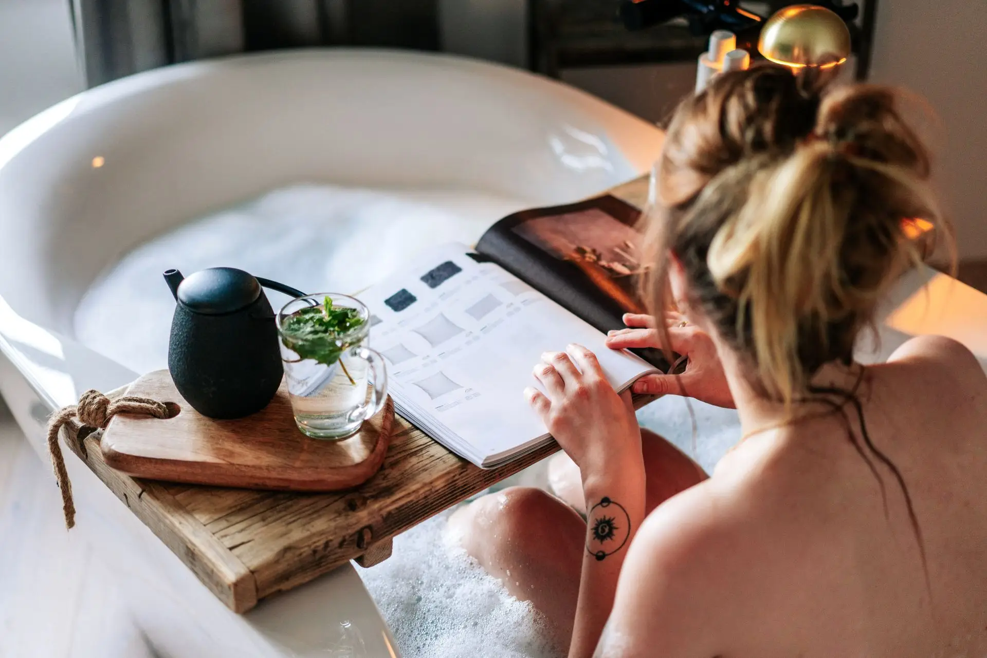 Frau entspannt in einer Badewanne, liest ein Buch, mit einem Teeglas und einer Kanne auf einem Holzbrett.