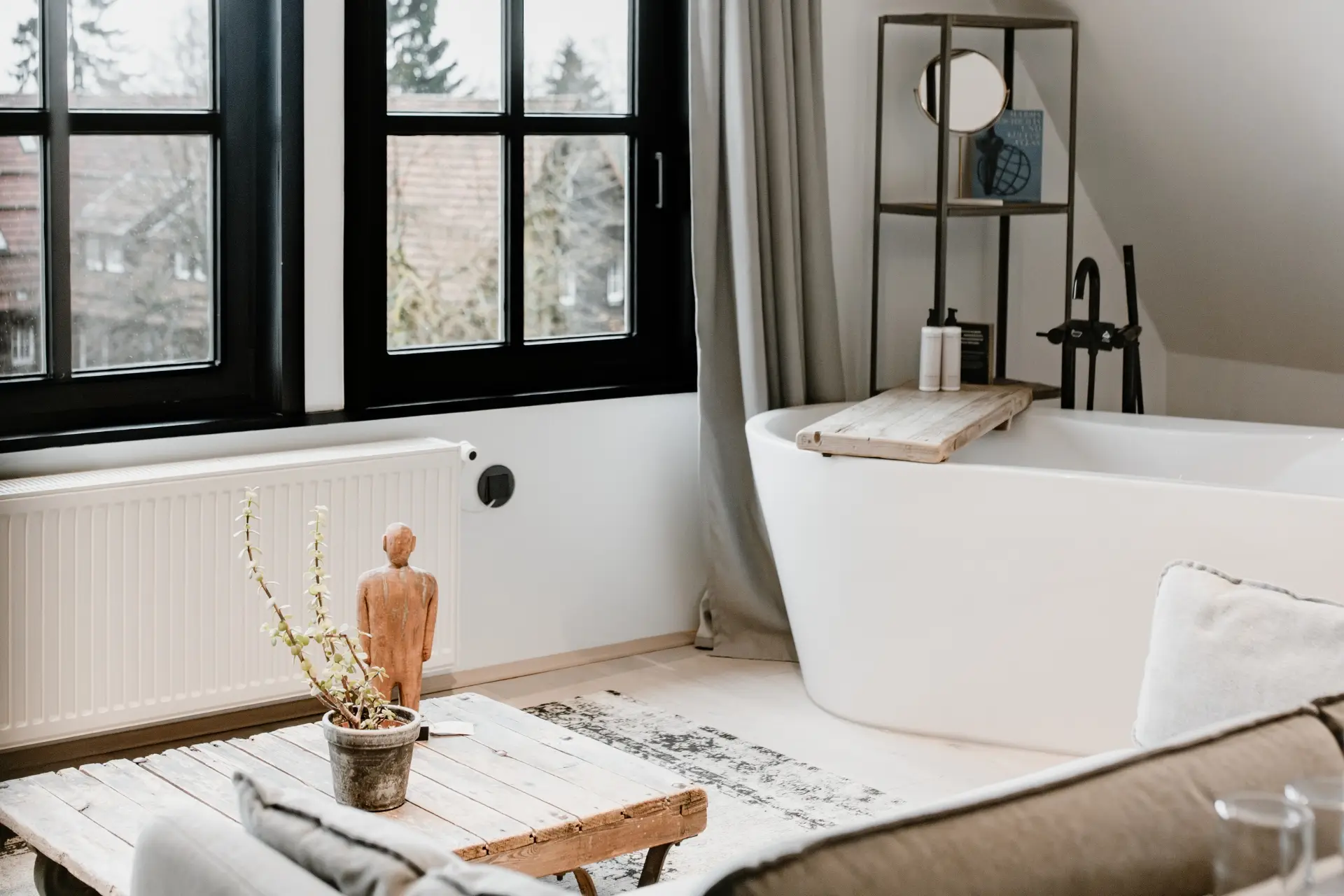 Helles Badezimmer mit freistehender Badewanne, Holzregal, Pflanzen und Blick auf die Fenster mit Vorhängen.