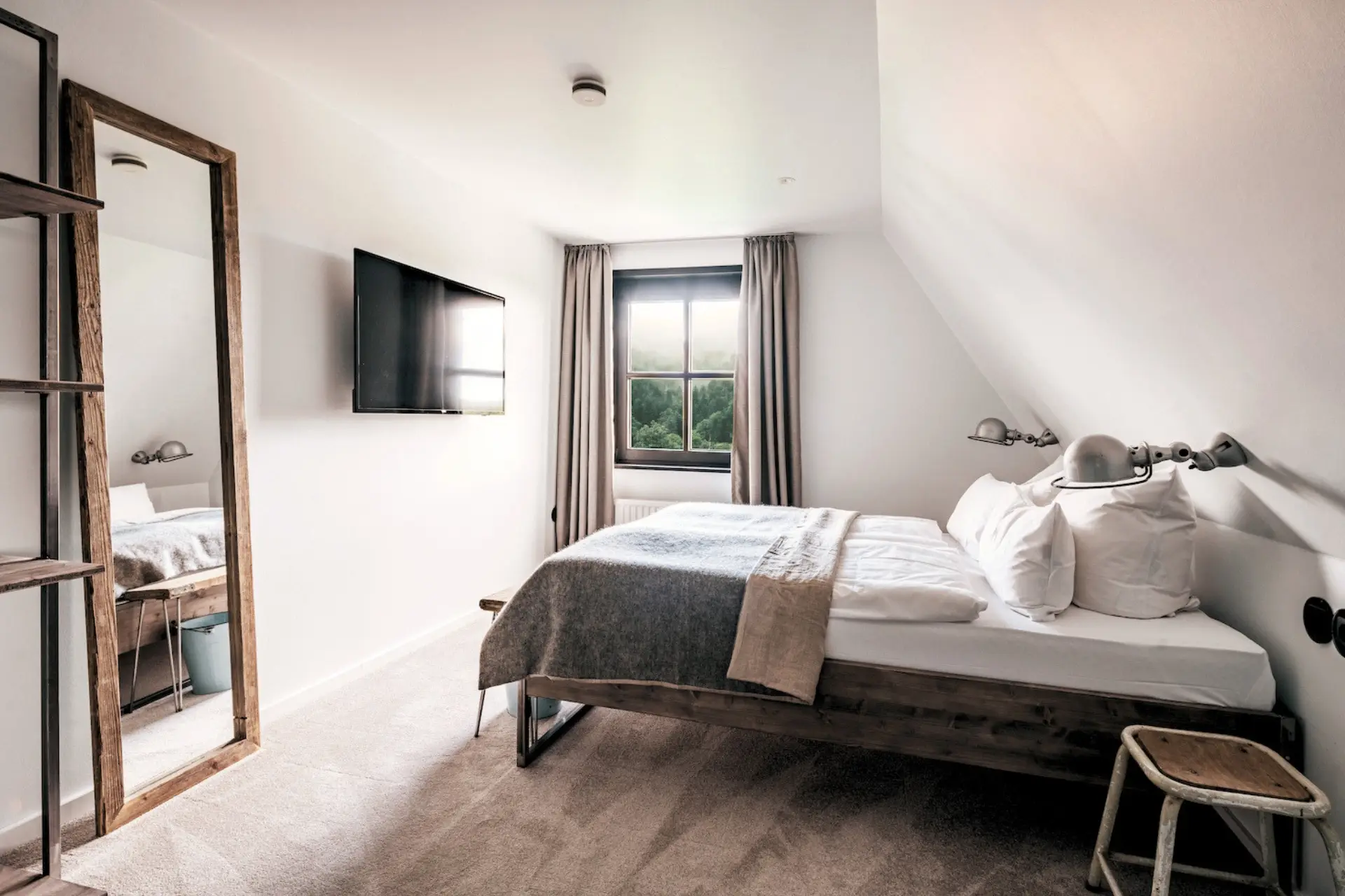 Minimalistisches Schlafzimmer mit Doppelbett, Fernseher, Wandspiegel und Verandafenster, beige Gardinen und warmen Lichtverhältnissen.