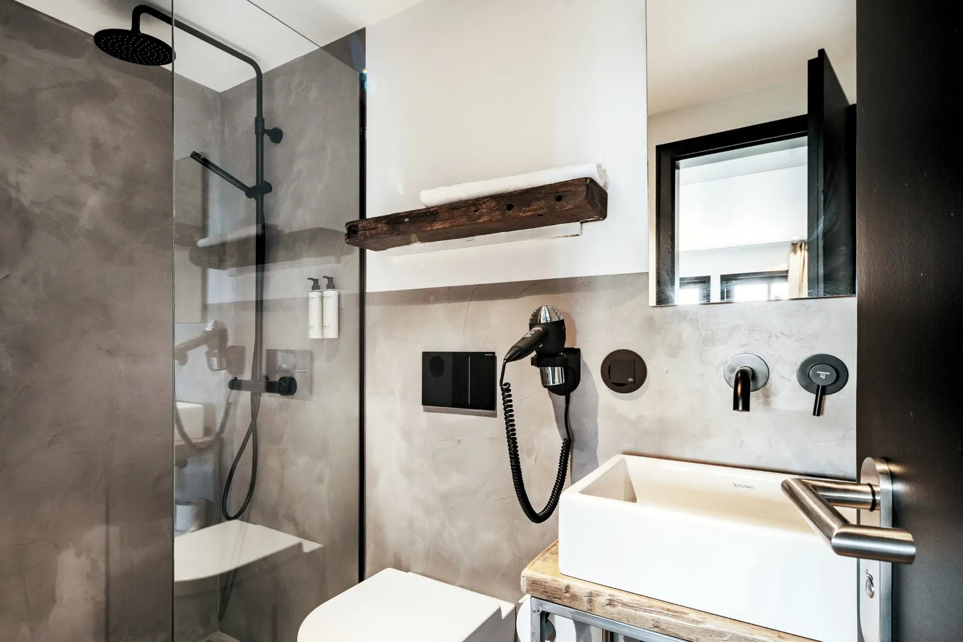 Modernes Badezimmer mit Dusche, Waschbecken aus Keramik, Wandregal aus Holz, Fön und grauen Wänden.