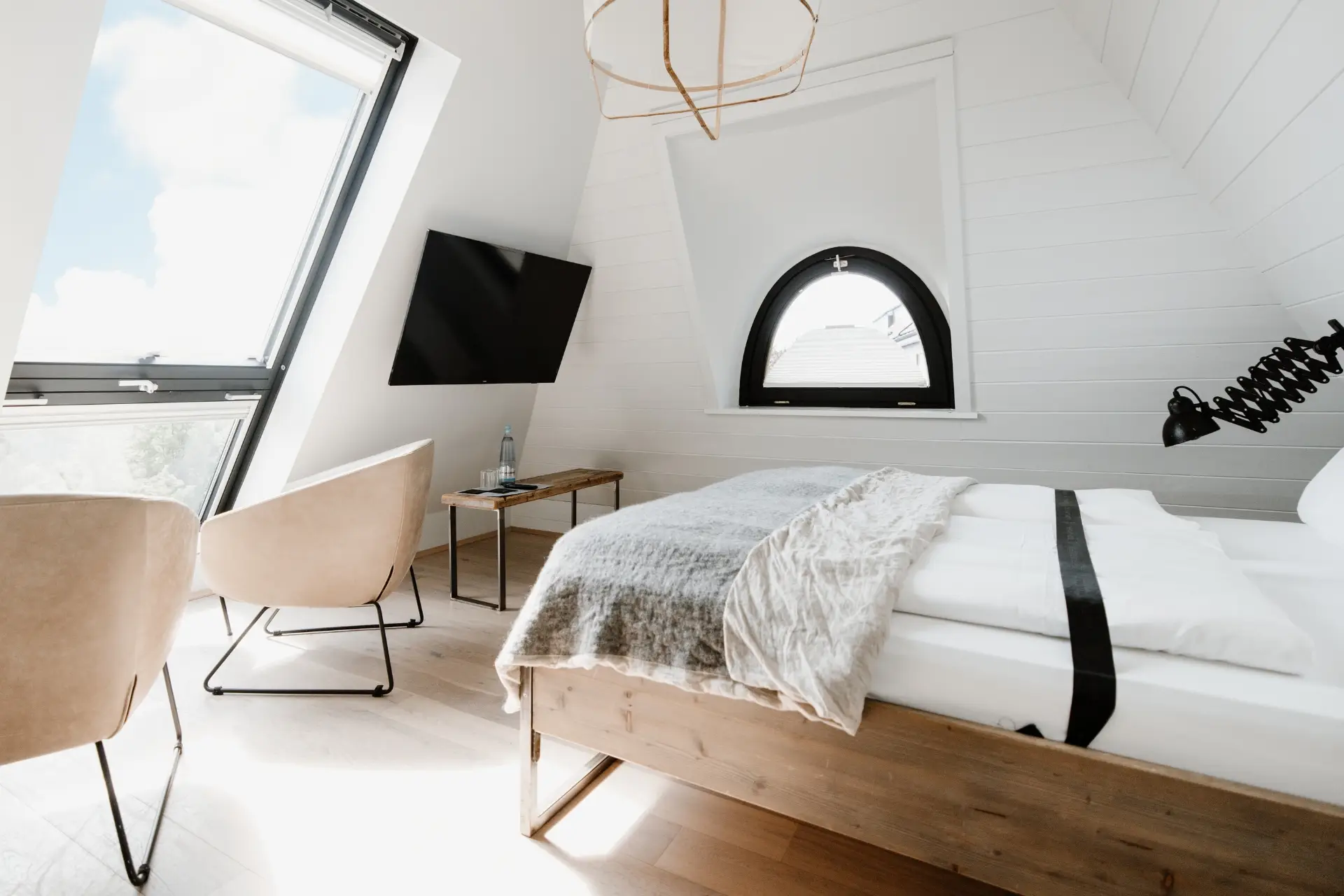 Helles Schlafzimmer mit Schrägdach, zwei Sessel, Tisch, TV und großem Fenster mit Blick auf den Himmel. Gemütliche Atmosphäre.