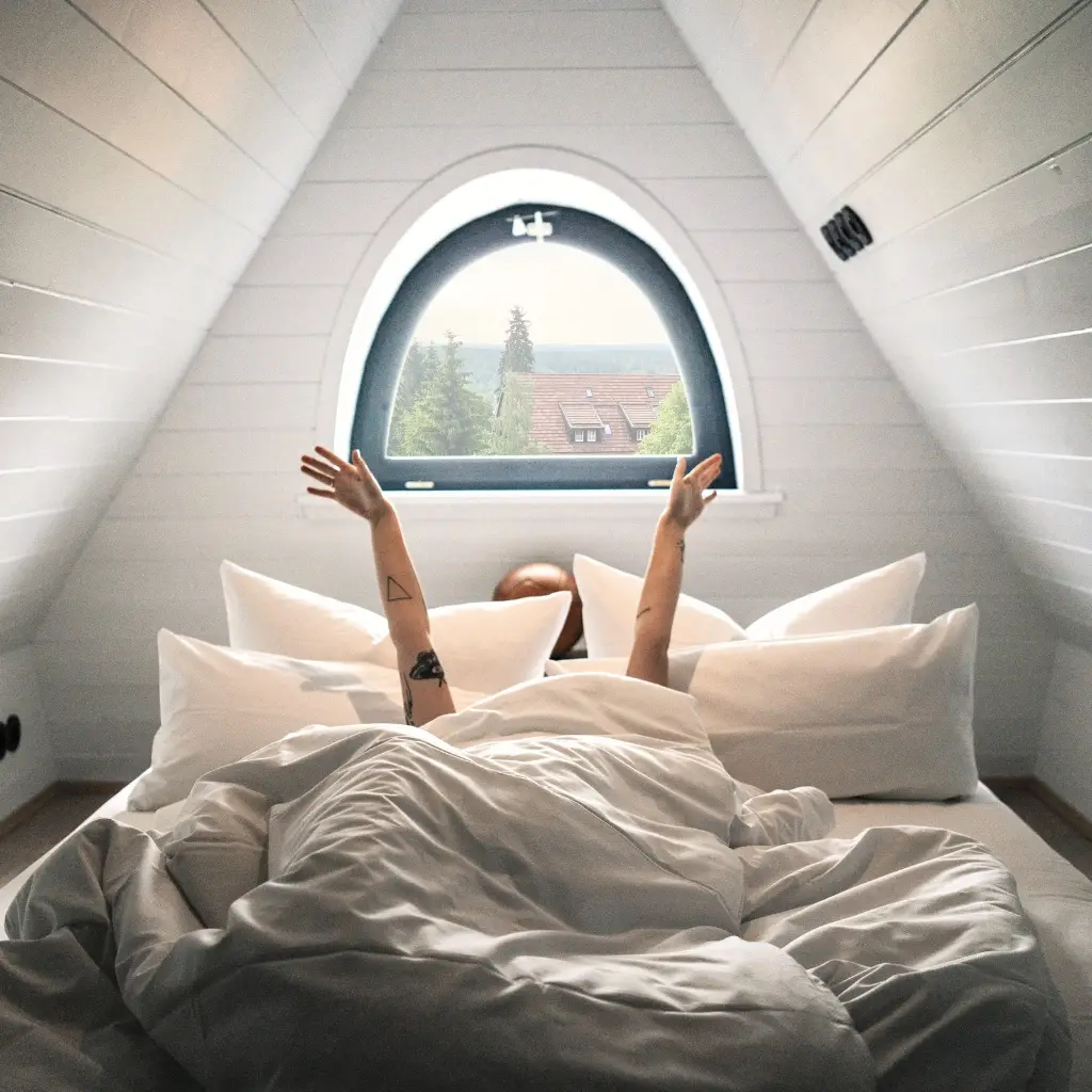 Person im Bett mit erhobenen Armen, umgeben von Kissen, Fenster mit Blick auf Bäume und Dächer im Hintergrund.