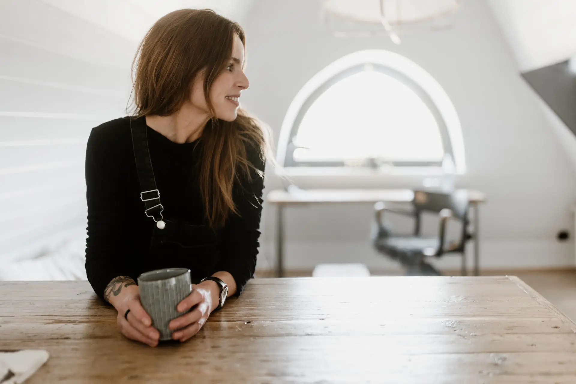 Frau mit langen Haaren sitzt am Tisch und hält eine Tasse, im Hintergrund ein heller Raum mit Fenster und Stuhl.