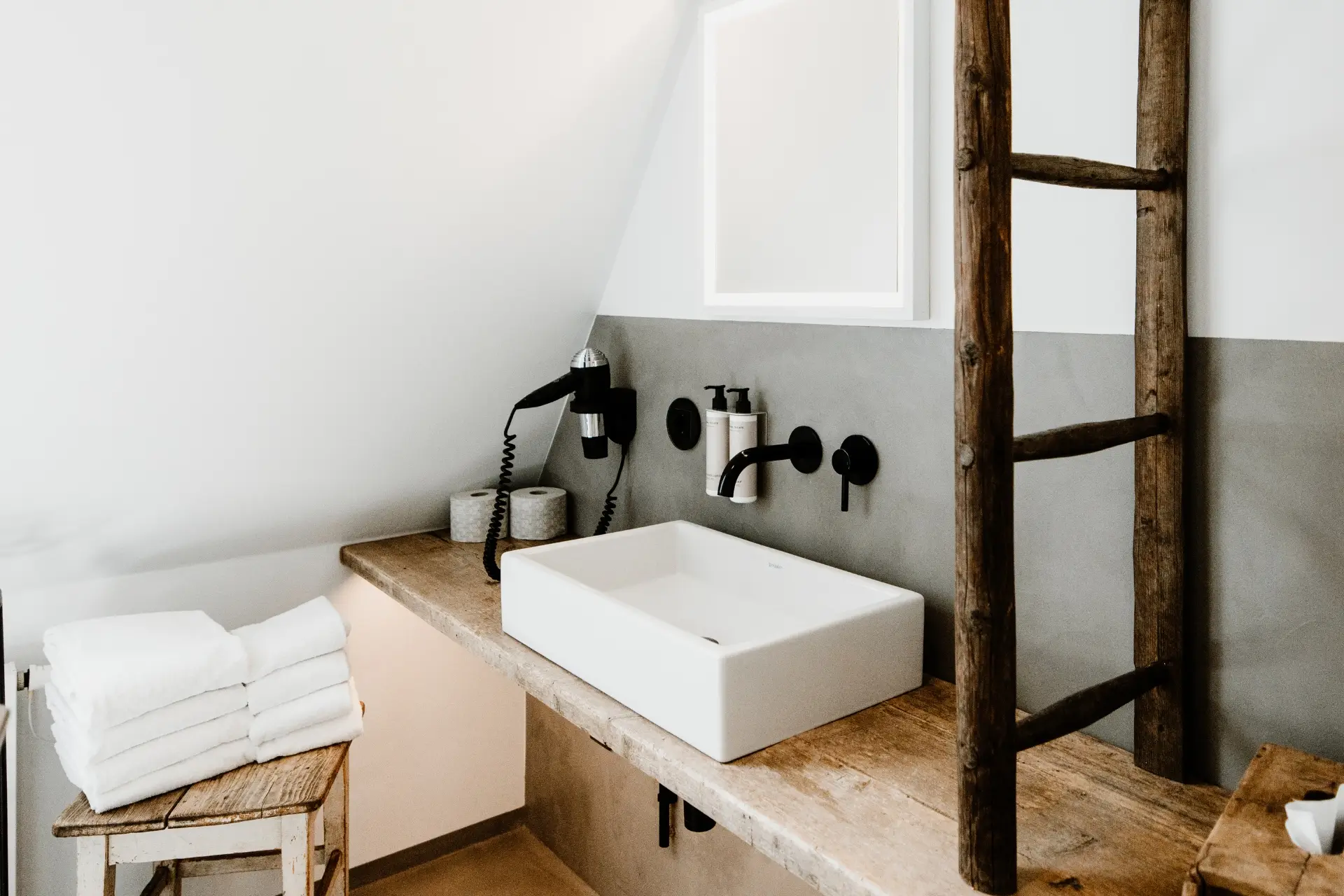 Modernes Badezimmer mit weißem Waschbecken, Holzregal, Handtuchstapel und Haartrockner an der Wand. Minimalistische Gestaltung.