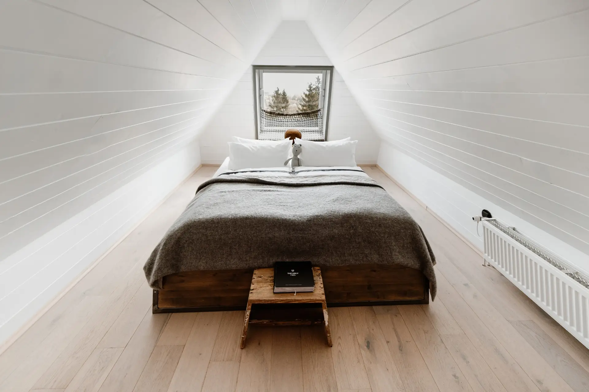 Helles Schlafzimmer im Dachgeschoss mit einem Bett, weißen Wänden und einem Fenster, das auf einen Waldblick zeigt.