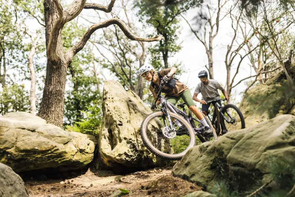 Zwei Radfahrer fahren über felsige, bewaldete Strecke; einer springt über einen großen Felsen, der andere folgt.