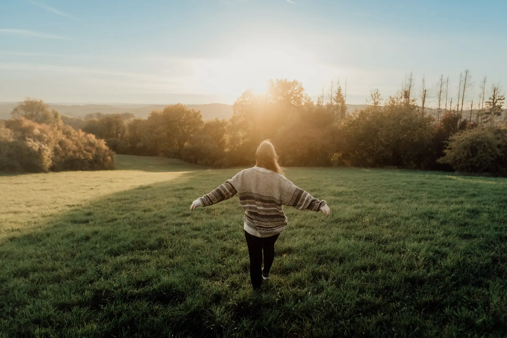 Eine Person mit langem Haar trägt einen Pullover und steht auf einer Wiese, während die Sonne aufgeht.