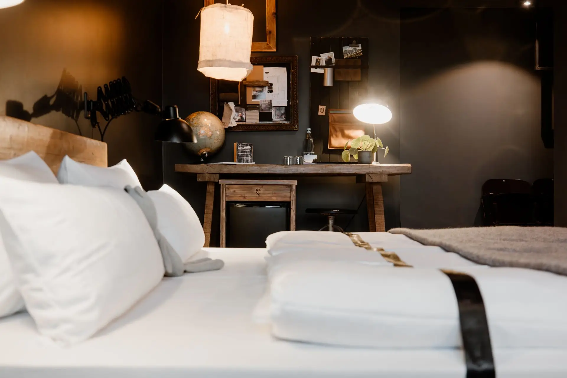 Gemütliches Boutique-Zimmer mit Bett, Holzschreibtisch, Globus und Pflanzen. Sanfte Beleuchtung und dunkle Wände schaffen eine einladende Atmosphäre.
