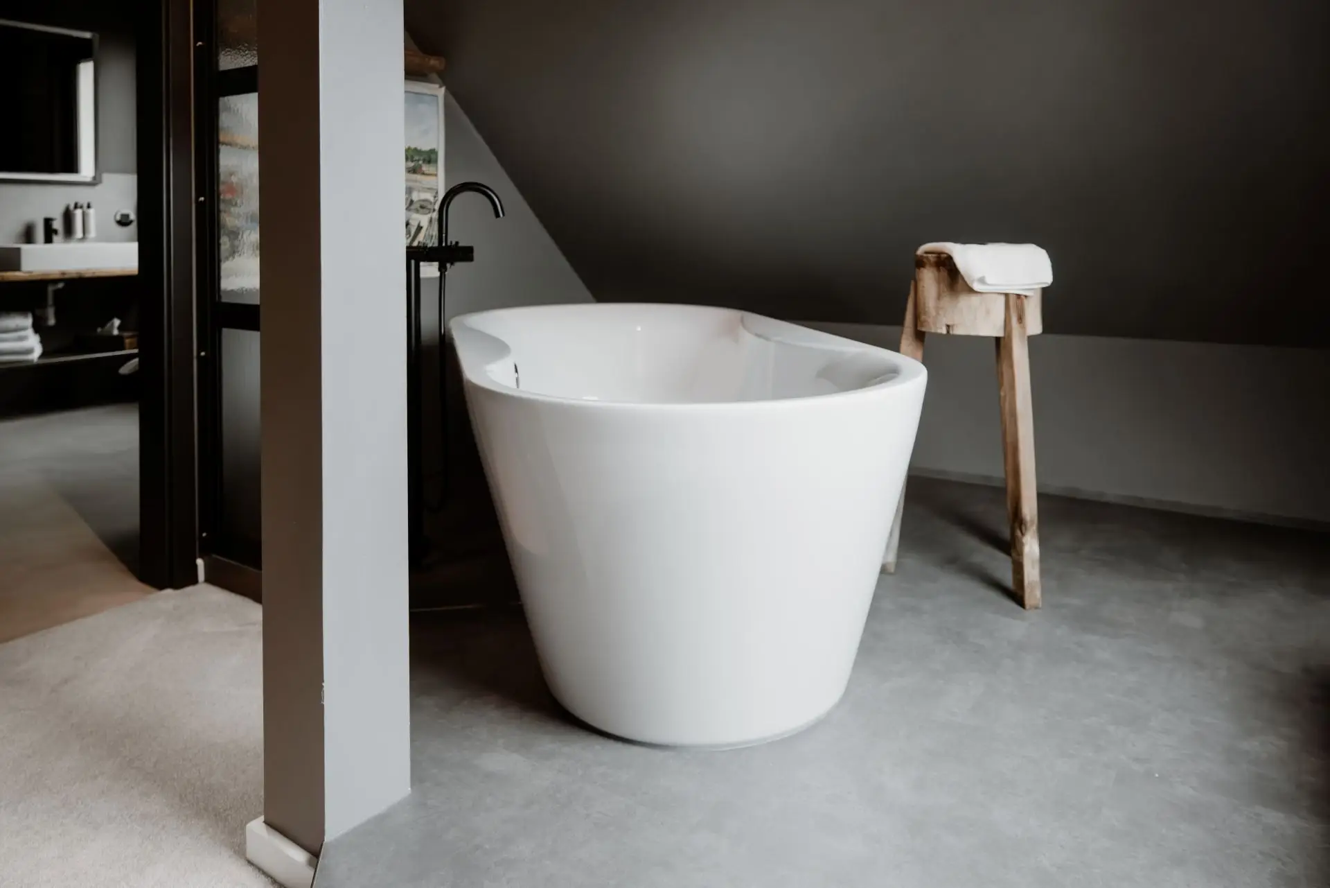 Weiße, freistehende Badewanne in einem modernen Bad mit grauen Wänden und einem Holzstuhl mit Handtuch.