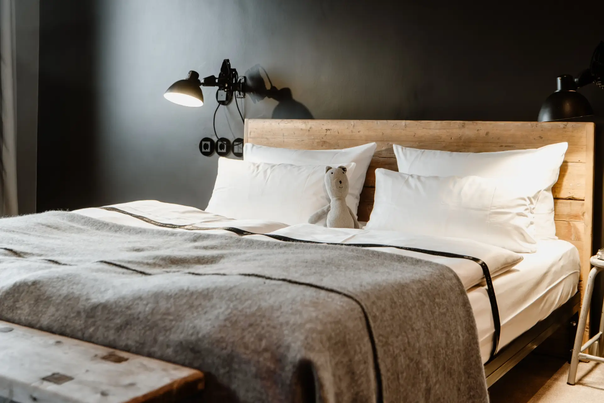 Doppelbett mit weißen Kissen und grauer Decke, neben einem Stofftier und Wandleuchten in einem modernen Schlafzimmer.