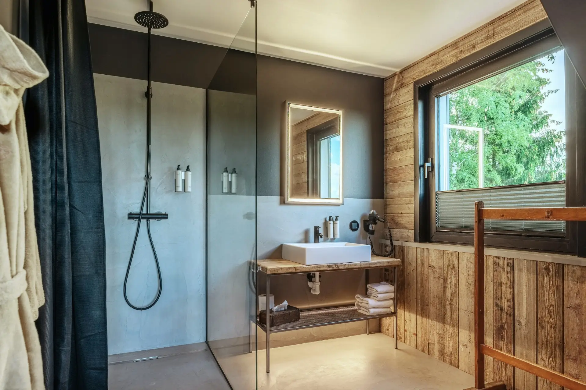 Modernes Badezimmer mit Dusche, Holzwand, Waschbecken und Fenster, das Tageslicht hereinlässt. Handtücher und Pflegeprodukte sind sichtbar.