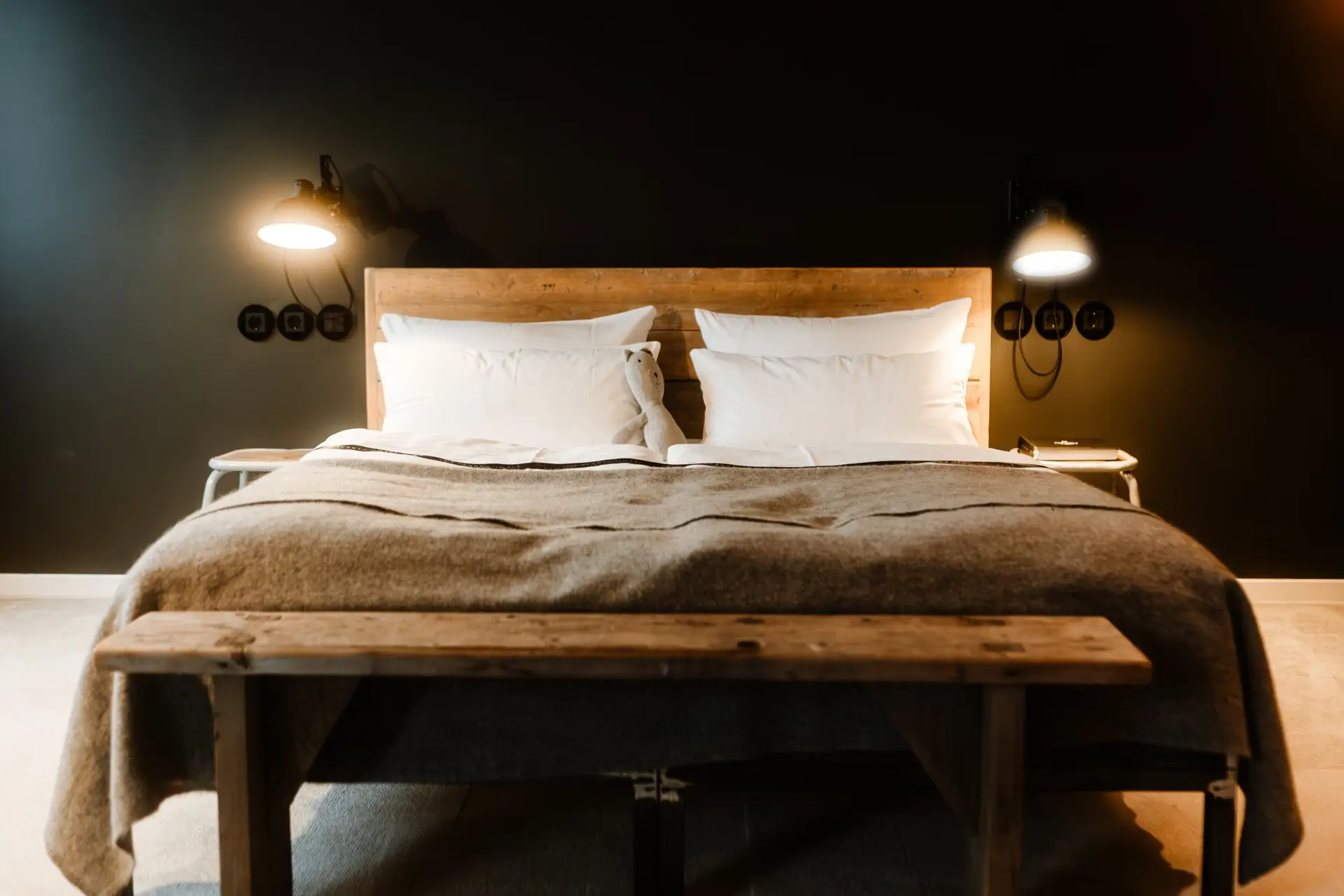 Doppelbett mit weißen Kissen und grauer Decke, Holzbank davor, an der Wand eine Leselampe und Steckdosen. Dunkle Wände.