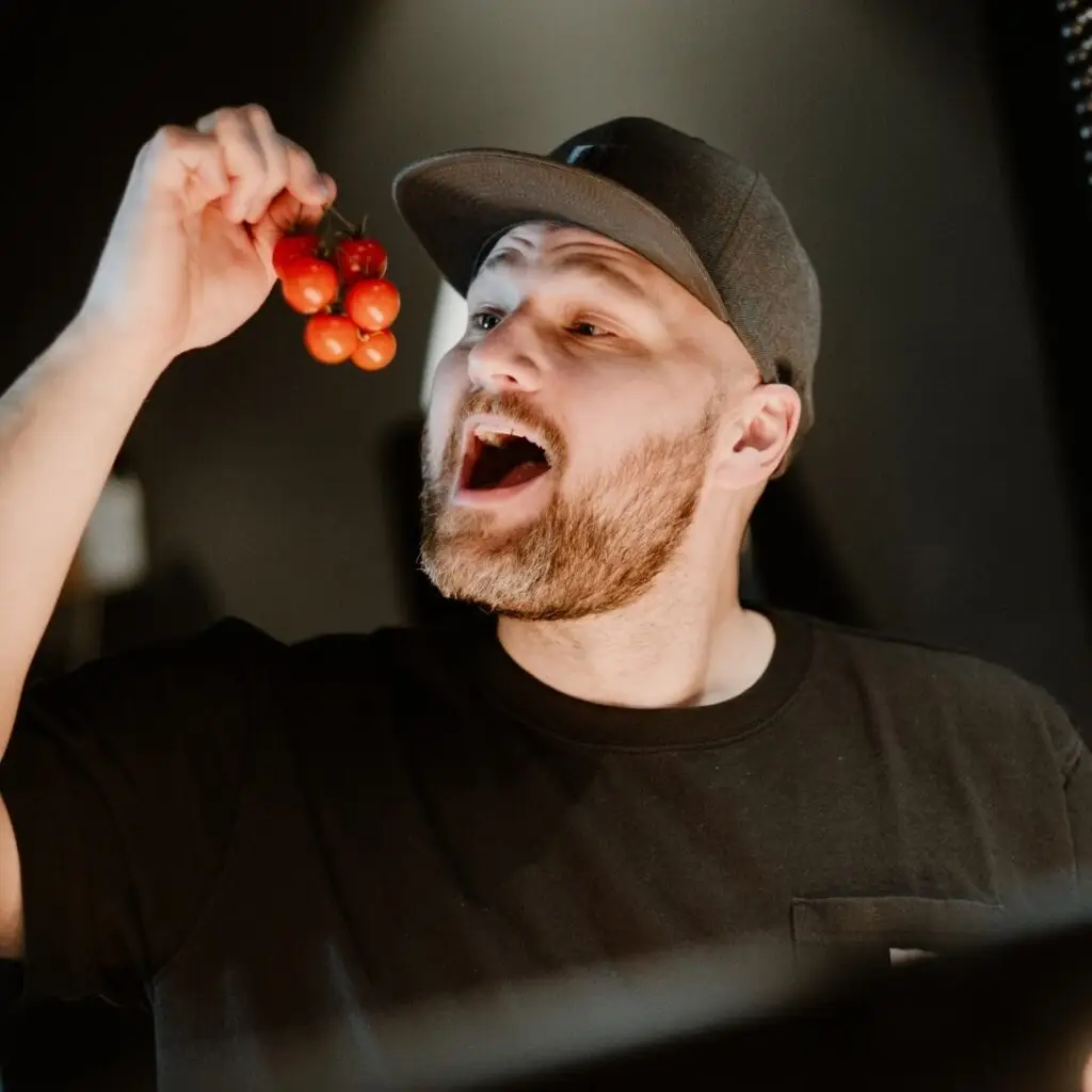 Mann mit Bart und Baseballkappe hält eine Traube Kirschtomaten hoch und lächelt aufmerksam in die Kamera.
