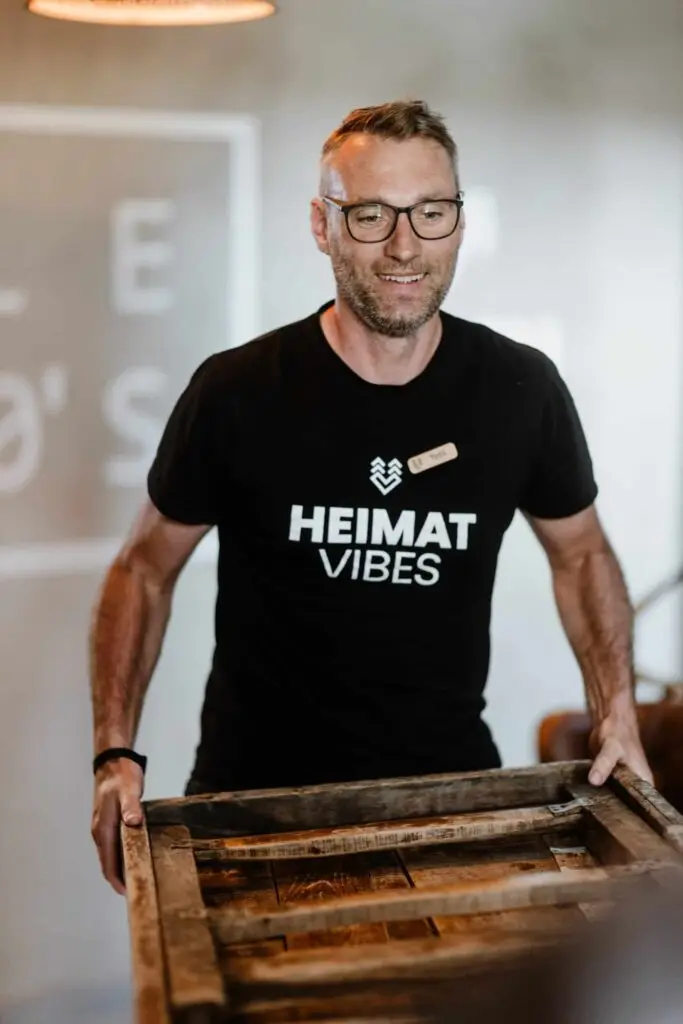 Mann mit Brille und T-Shirt mit der Aufschrift „HEIMAT VIBES“ trägt einen Holztisch in einem modernen Innenraum.