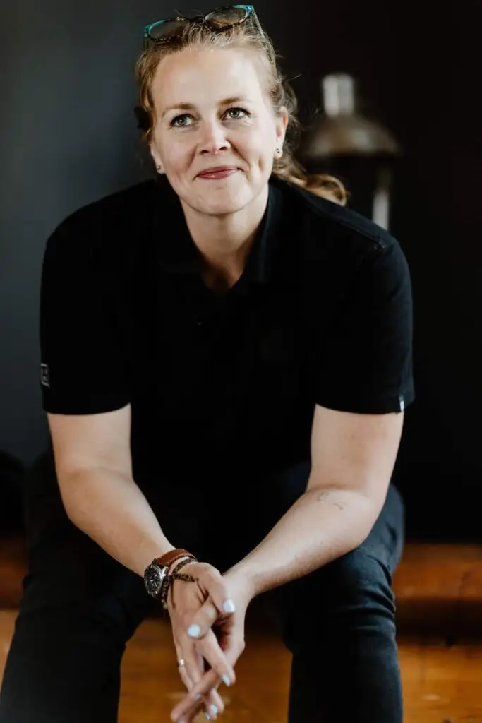 Portrait einer Frau mit lächelndem Gesicht, trägt schwarzes Poloshirt, sitzt mit gefalteten Händen in einem modernen Raum.