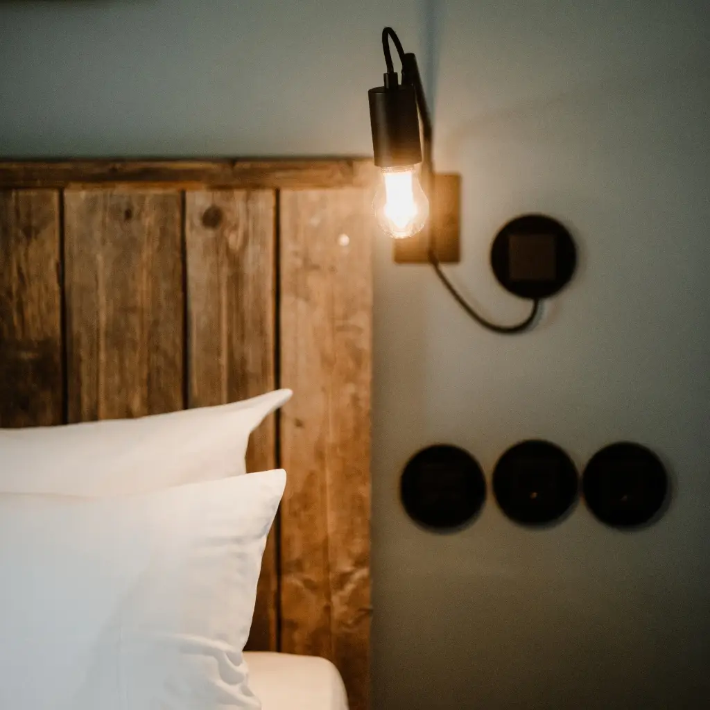 Beleuchtetes Bett mit Kissen vor einer Holzpaneelwand und Steckdosen in einem modernen Schlafzimmer.
