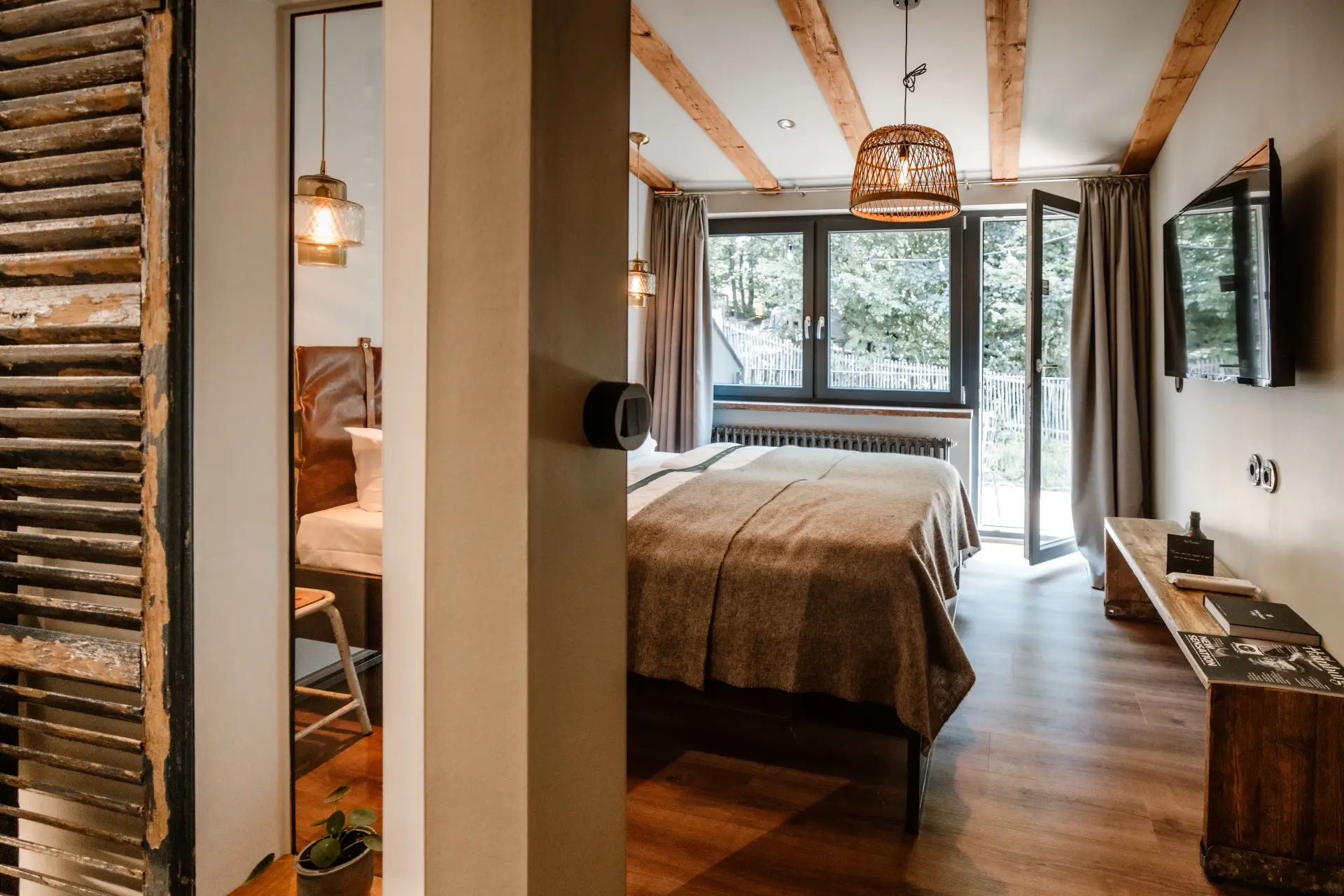 Helles Zimmer mit Holzbalken, offenem Fenster, Doppelbett, TV und einem glänzenden Licht. Im Hintergrund eine Pflanzenkanne.
