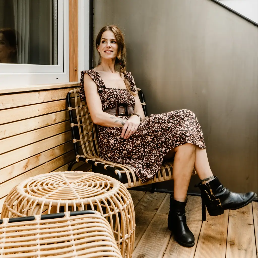 Eine Frau in einem blumigen Kleid sitzt auf einem Designerstuhl auf einer Holzterrasse, lächelnd in die Kamera.
